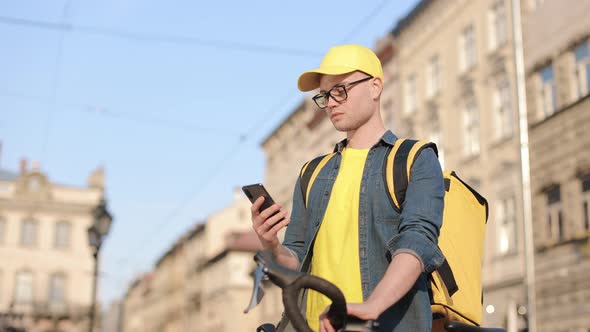 Portrait of a Happy Delivery Man Who is Sitting on a Bicycle and Texting on the Smartphone