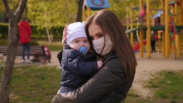 A Mother in a Medical Mask Hugs Her Little Son in a Park on a Crowded Playground