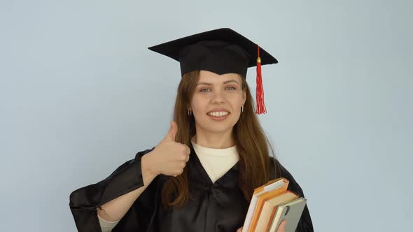 A Young Caucasian Woman in a Black Robe and a Master's Hat Stands Straight Holding Textbooks and