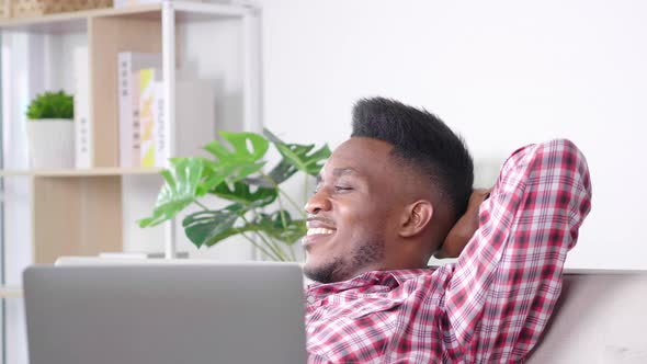 Happy African man feels relaxing on sofa in break time from work, putting laptop