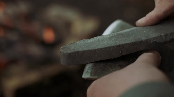 Close-up Of Casual Strong Man Works with Ax and Sharpening Stone Near Warm Bonfire in Camp in Wild