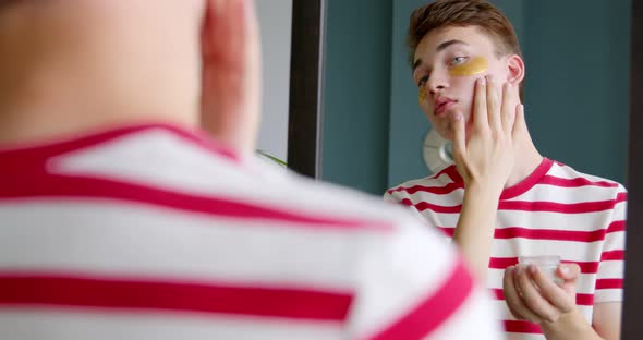 Young man applying moisturizing cream on face look in the mirror. Home skin care