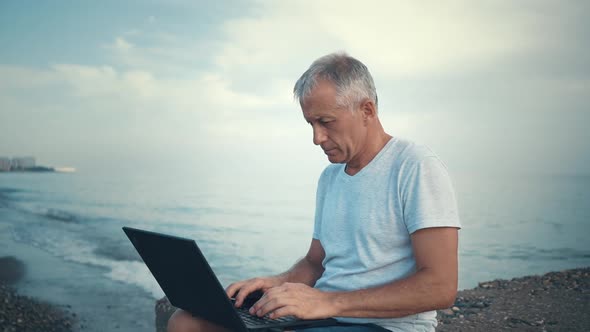 An Elderly Whitehaired Man Sits on the Beach with a Laptop and Studies the Information