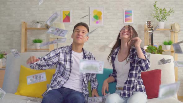 Young Asian Couple Sitting on Sofa Catching Paper Banknotes Slow Mo