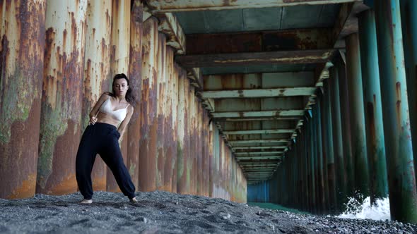 a Barefoot Woman Dances Under the Pillars of the Bridge Against the Background of the Incoming Waves