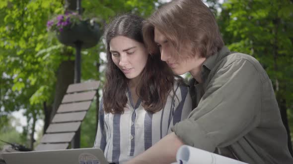 The Male and Female Students in Casual Clothes Sitting at the Bench, Girl Holding Laptop and Boy Has