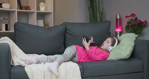Little Girl in Wireless Headphones Chilling Laughing on Sofa Listening to Favorite Music