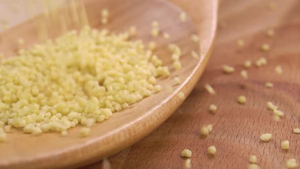 Raw uncooked couscous falls into an empty wooden spoon on a wood surface in slow motion