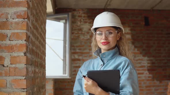 A Female Architect or Bricklayer Stands in a Newly Built House with Untreated Walls with a Tablet in
