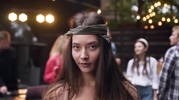 Young Woman in a Bandanna with Long Hair Posing to the Camera while Her Friends Dancing