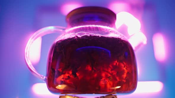 Transparent Teapot with Tea Leaves Boils on Gas Burner Against Neon Backlight