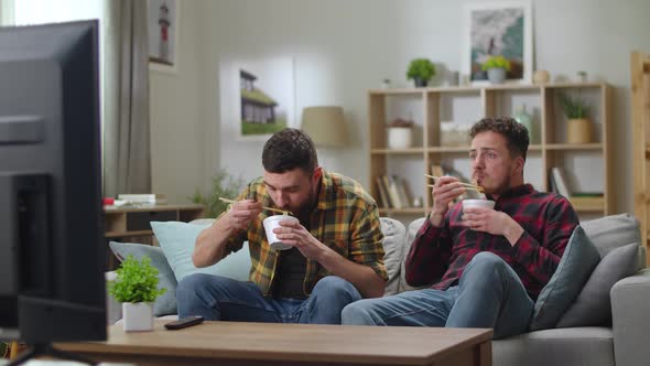 Two Young Cute Men in the Living Room on the Couch Watch TV and Eat Food From Delivery