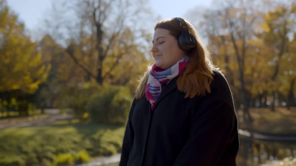 Young Happy Overweight Woman in Headphones Listening To Music and Dancing During Walk in Fall Park