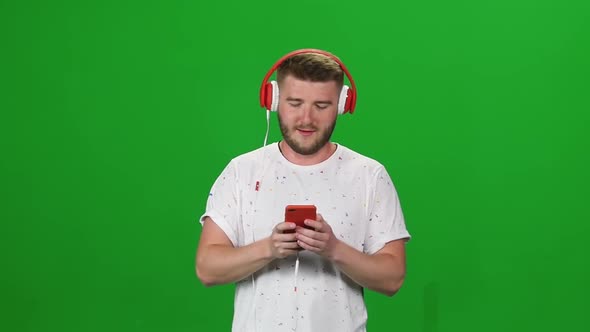Portrait Bearded Guy in Red Headphones Walking and Listening Music Vie Phone at Greenscreen. Front