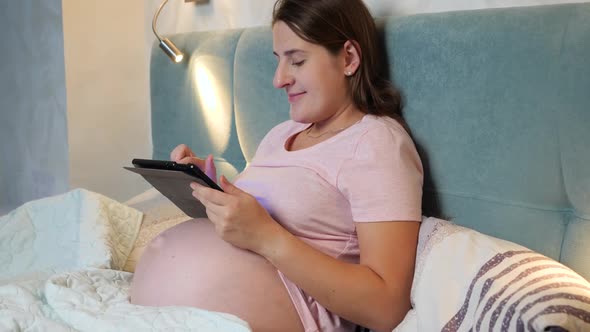 Happy Pregnant Woman in Pajamas Using Tablet Computer in Bed Before Going To Sleep at Night