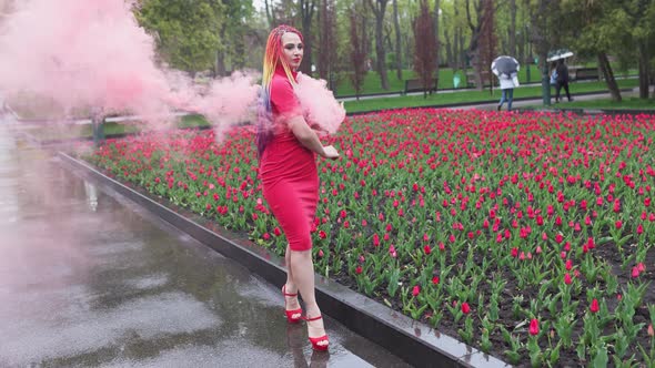 A Girl with Makeup with Rainbow Braids in Red Dress Posing in Red Smoke Against the Background of a