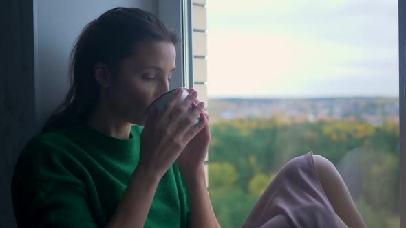 Woman in a Green Knitted Sweater Sits on the Window with an Iron Mug