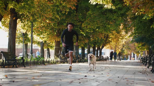 Young African American Black Man Running with His White Labrador Dog Through the Park in the City at
