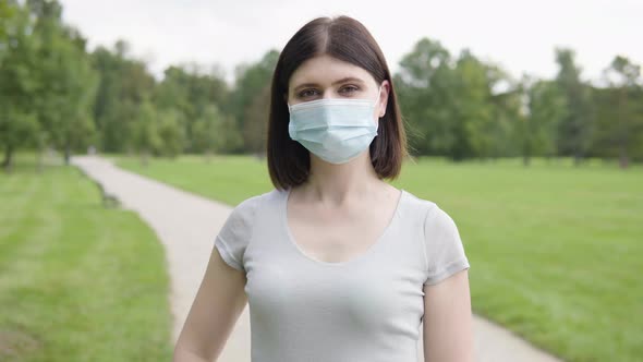 A Young Caucasian Woman in a Face Mask Shows a Thumb Up to the Camera and Nods on a Pathway in Park