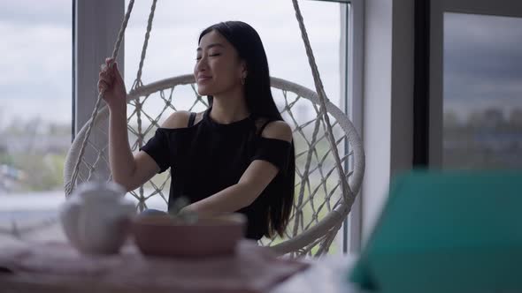 Happy Carefree Relaxed Asian Woman Spinning Sitting in Hanging Chair at Home