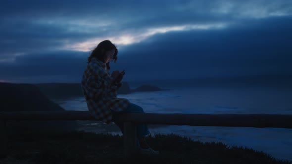 Young Girl Sits on Sea Shore Using Phone at Night