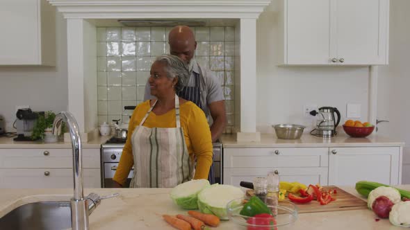 African american senior man tying apron from back to his wife in the kitchen at home