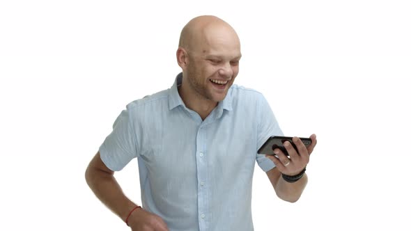 Handsome Bald Bearded Guy with Blue Shirt Looking at Mobile Phone and Watching Video Laughing at