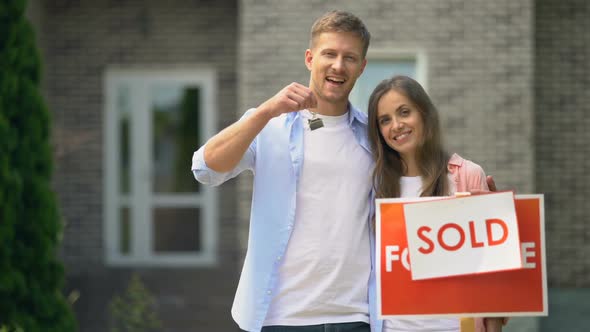 Happy Family Couple Smiling, Holding House Keys Standing Near Sold Signboard