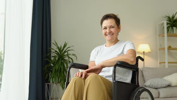 Portrait of Mature Smiling Woman Sitting in Wheelchair at Home and Looking at Camera