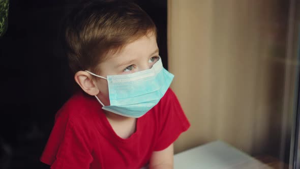 Young Boy in a Medical Mask Looks Out the Window. Self-isolation in Quarantine, Coronavirus, Covid