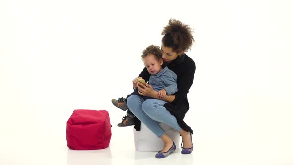 African American Mother Is Sitting with Her Baby, Holding a Phone. White Background. Slow Motion