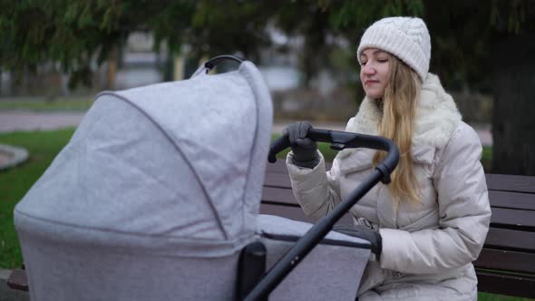 Young Mother with Newborn Child Outdoor. She Sitting on the Bench with Baby Sleeping in Pram