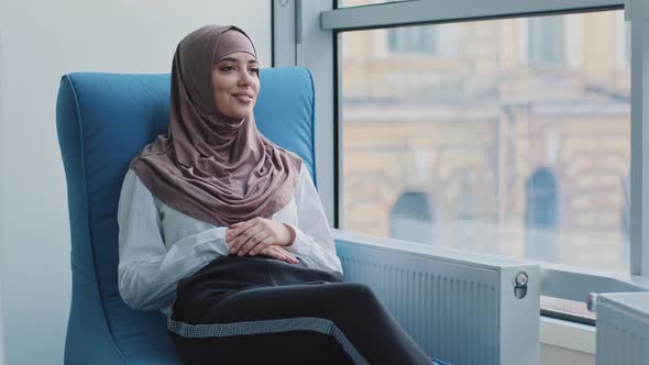 Smiling Dreamy Arabic Girl in Hijab Looking Out Window Sitting in Armchair Waiting for Exam or