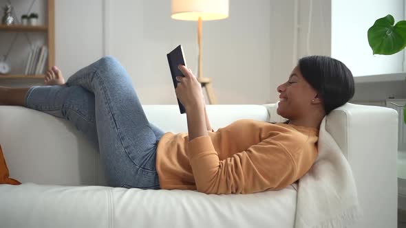 African Woman Who is Lying on Sofa in Home Interior Holds Book in Hand Spbd