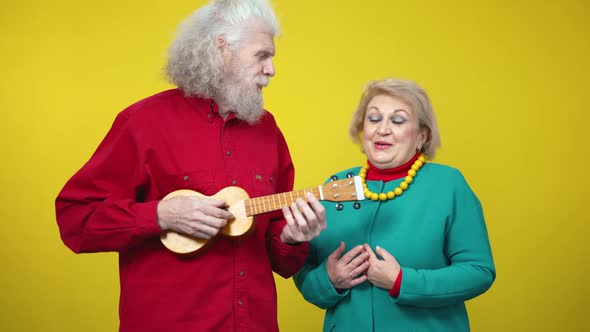 Positive Senior Husband Playing Ukulele and Singing Serenade for Wife at Yellow Background