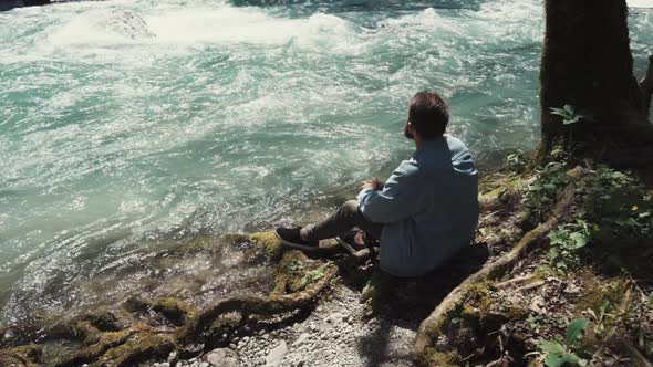 a Young Man Sits Near a Fast Stream of a River and Looks at the Running Water