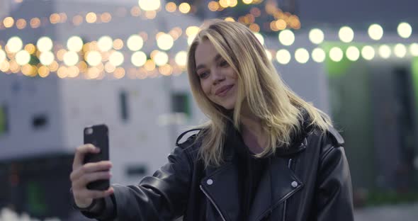 Young Girl Holds a Smartphone in Her Hand and Takes a Selfie Outside in a Modern Residential Area