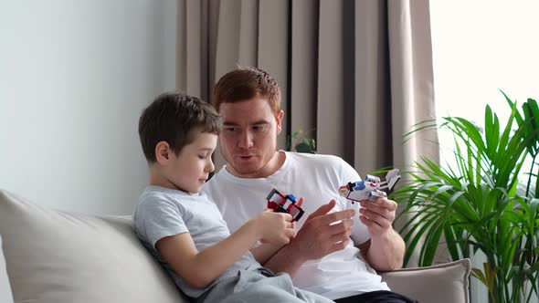 Father with Son Sitting on Sofa Indoors and Playing with Plastic Robots