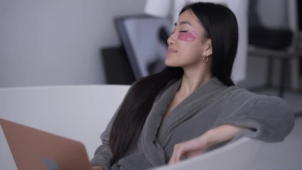 Relaxed Happy Asian Woman with Eye Patches Sitting in Bathtub in Bathrobe Smiling