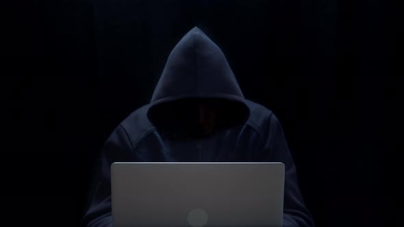 The hacker uses a laptop. A hooded mysterious man with an invisible face in a dark room.