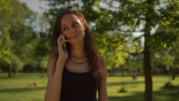 Smiling Young Lady Standing at the Park Outdoors Talking By Phone