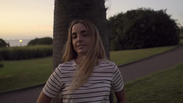 Slow motion shot of young blond woman leaning on tree trunk in park