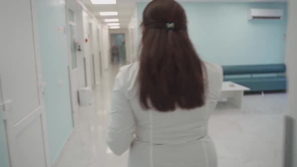 Woman Doctor in White Coat Walks Along Corridor of Clinic with Patient Card or Documents in Hand and