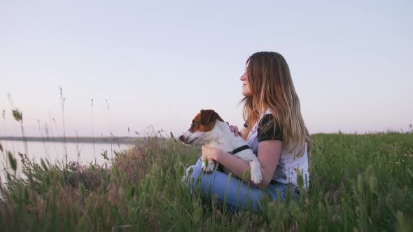 Young Happy Woman and Het Little Dog Sitting with Flying Kite on a Glade at Sunset