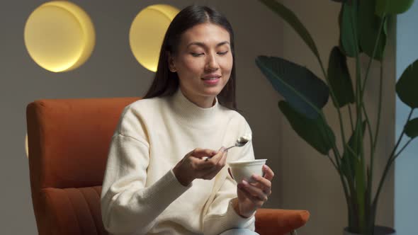 Cute Young Woman Eating Yogurt Sitting at Home on a Chair