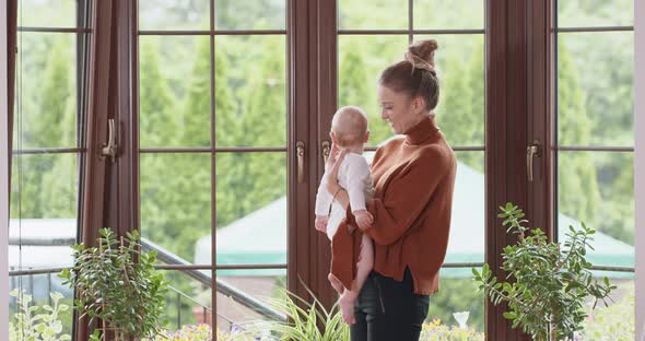 Young Mother Holds Her Adorable Baby and Stays with It in Front of the Window Kissing Baby Window