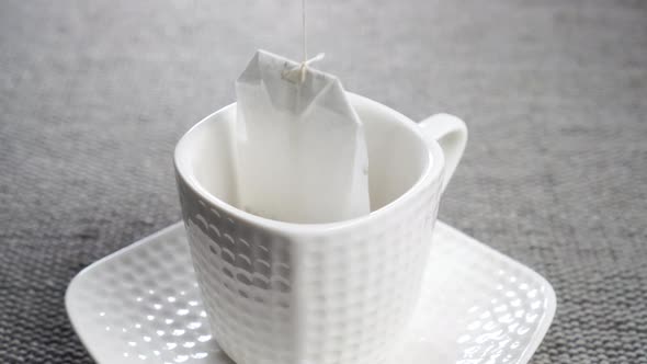 Dipping a tea bag into a empty white teacup with saucer on a gray textured gray mat