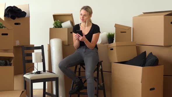 A Happy Moving Woman Sits on a Chair in an Empty Apartment and Works on a Smartphone with a Smile