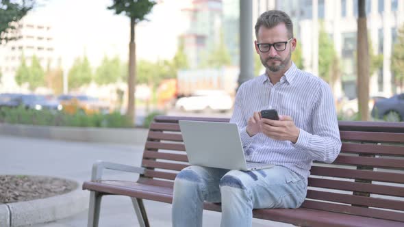 Young Adult Man Using Smartphone and Laptop While Sitting Outdoor on Bench