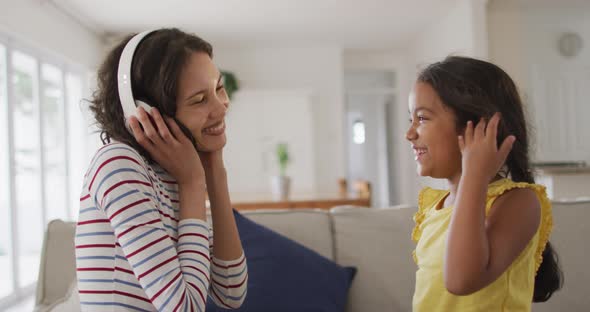 Happy hispanic mother wearing headphones listening to music and daughter sitting on sofa